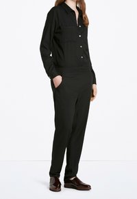 Zwarte jumpsuit met lange mouwen, twee borstzakken, knoopsluiting en taps toelopende benen. Gemaakt van een soepele stof met een overall stijlvolle afwerking.