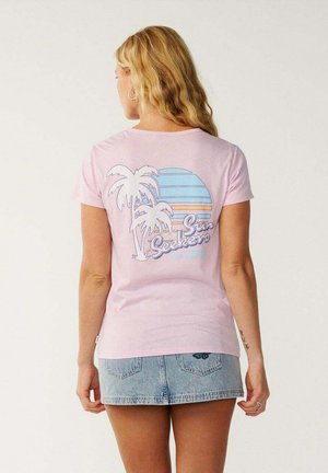 Femme aux longs cheveux blonds portant un t-shirt rose "Sun Seekers" avec un graphisme de palmier et de coucher de soleil, et une mini-jupe en jean bleu clair.