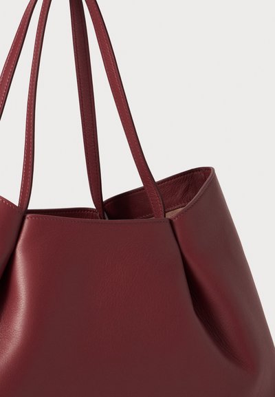 Geantă tote din piele burgundă, cu două mânere de umăr, textură moale și formă rotunjită. Dispune de un design minimalist, fără accesorii vizibile.