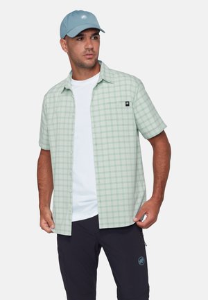 Uomo che indossa una camicia a maniche corte a quadri verde chiaro con bottoni, maglietta bianca, pantaloncini scuri e cappellino blu su uno sfondo semplice.