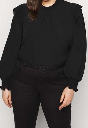 Donna che indossa una camicetta nera a maniche lunghe con spalle e polsini arricciati, abbinata a pantaloni neri a vita alta.