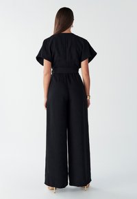 Črn kombinezon s kratkimi, širokimi rokavi in pasom. Razširjene hlačnice sežejo do gležnja, z gladko teksturo in minimalističnimi detajli.