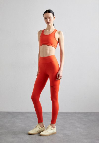 Bright orange athletic crop top and leggings made of stretchy material, featuring flat seams and minimalist design. Worn with beige sneakers.