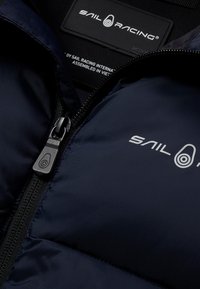 Marineblaue Steppjacke mit glatter Oberfläche. Mit einem Frontreißverschluss und dem weißen "SAIL RACING"-Logo auf der linken Seite. Tags innen sichtbar.
