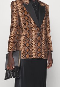 Blazer à motif serpent marron et noir avec des revers en satin, associé à une jupe noire et une pochette noire ornée de franges en perles.