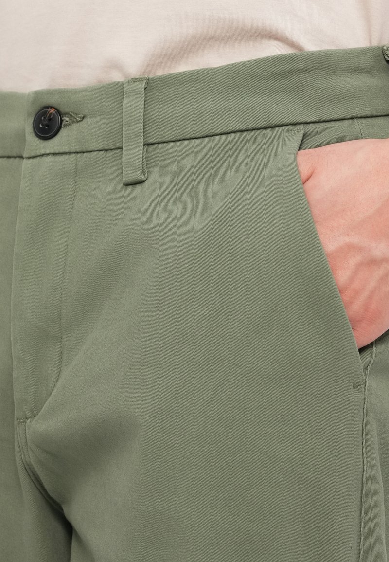 Primer plano de una mano en el bolsillo delantero de unos pantalones verde oliva con detalles de botón y trabilla, combinados con una blusa beige.