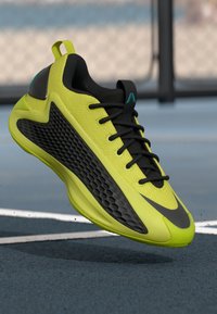 Sapatilha desportiva em amarelo brilhante e preto com atacadores pretos, suspensa acima de um campo de ténis azul com linhas brancas e uma vedação de malha ao fundo.