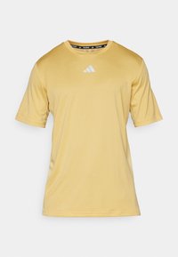 Žluté triko s krátkým rukávem značky Adidas na trénink s kulatým výstřihem a bílým logem Adidas umístěným na hrudi na jednobarevném pozadí.