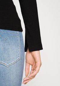 Die rechte Hand entspannt neben der Tasche einer hellblauen Jeans, während ein schwarzes Langarmoberteil mit einem kleinen Seitenschlitz am Ärmel getragen wird.