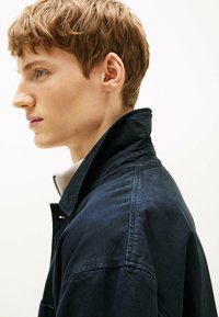 Marineblauwe jas met een hoge kraag, voorzien van een rits en een gestructureerde stof. Model draagt een licht shirt eronder, getoond in profiel.