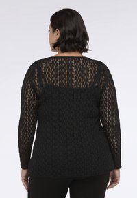 Žena s krátkými tmavými vlasy, oblečená v černém krajkovém topu s dlouhým rukávem přes černý tílko, stojící na jednobarevném pozadí.