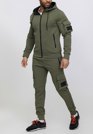 Survêtement à capuche vert olive en tissu texturé. Présente un devant zippé, des poches latérales et une poche style cargo sur le pantalon. Accents noirs.