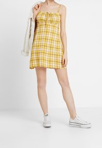 Keltainen ruudullinen mekko, jossa on säädettävät olkaimet ja solmittava etuosa. Yhdistettynä valkoisiin tennareihin, mikä korostaa leikkisää ja rentoa tyyliä. Tasainen helma.