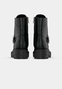 Botas negras hasta el tobillo, hechas de cuero liso, con cremallera lateral, suela gruesa y resistente, y una correa decorativa con detalle de hebilla metálica.