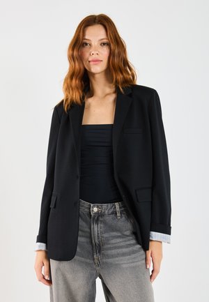 Blazer noir avec revers, fermeture à un bouton et détails de poche avant. Associé à un haut noir et un jean gris taille haute. Tissu en mélange de coton.