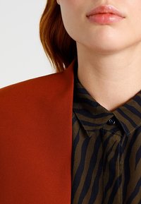 Blazer arancione con una texture liscia, abbinato a una camicia verde scuro con strisce nere verticali e colletto a punte.