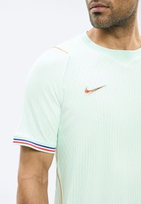 Άνδρας που φοράει ανοιχτό πράσινο αθλητικό μπλουζάκι Nike με πορτοκαλί διακοσμητική λωρίδα και κόκκινες, λευκές και μπλε ρίγες στο μανίκι, εμφανιζόμενος από τον ώμο έως το πηγούνι.