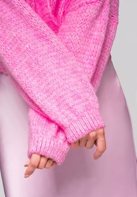 Chunky pletený svetr v jasně růžové barvě, který ukazuje texturovanou látku a žebrované manžety. Rukáv přesahuje hladký pastelově růžový pozadí.