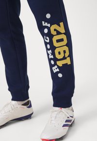 As calças de treino em azul-marinho apresentam gráficos em amarelo e branco, incluindo "1902" e "R.M.G.F.", combinadas com chuteiras de futebol brancas com acabamentos em azul.