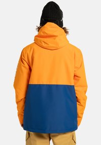 Osoba w dwukolorowej pomarańczowo-niebieskiej kurtce z kapturem i czarnym beanie z goglami, stojąca tyłem na jednolitym tle.