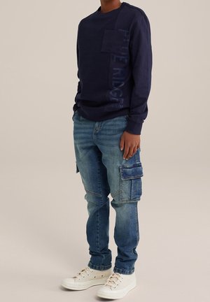 Marineblauw shirt met lange mouwen, een gestructureerde afwerking en een zijzak; blauwe cargobroek met meerdere zakken en een relaxte pasvorm; witte sneakers.