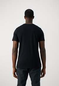 Camiseta negra de manga corta hecha de algodón, con cuello redondo y una textura suave. Persona de pie de espaldas al espectador.
