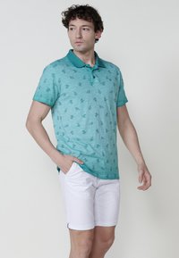 Türkiz színű, rövid ujjú polo póló, levélmintázattal, gallérral és két gombbal. Fehér sorttal viselik. Simább textúra.