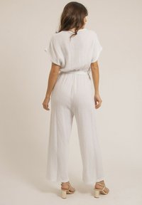Combinaison blanche et respirante en tissu texturé, dotée de manches courtes, d'une coupe décontractée et d'une taille nouée. Assortie à des talons à brides.