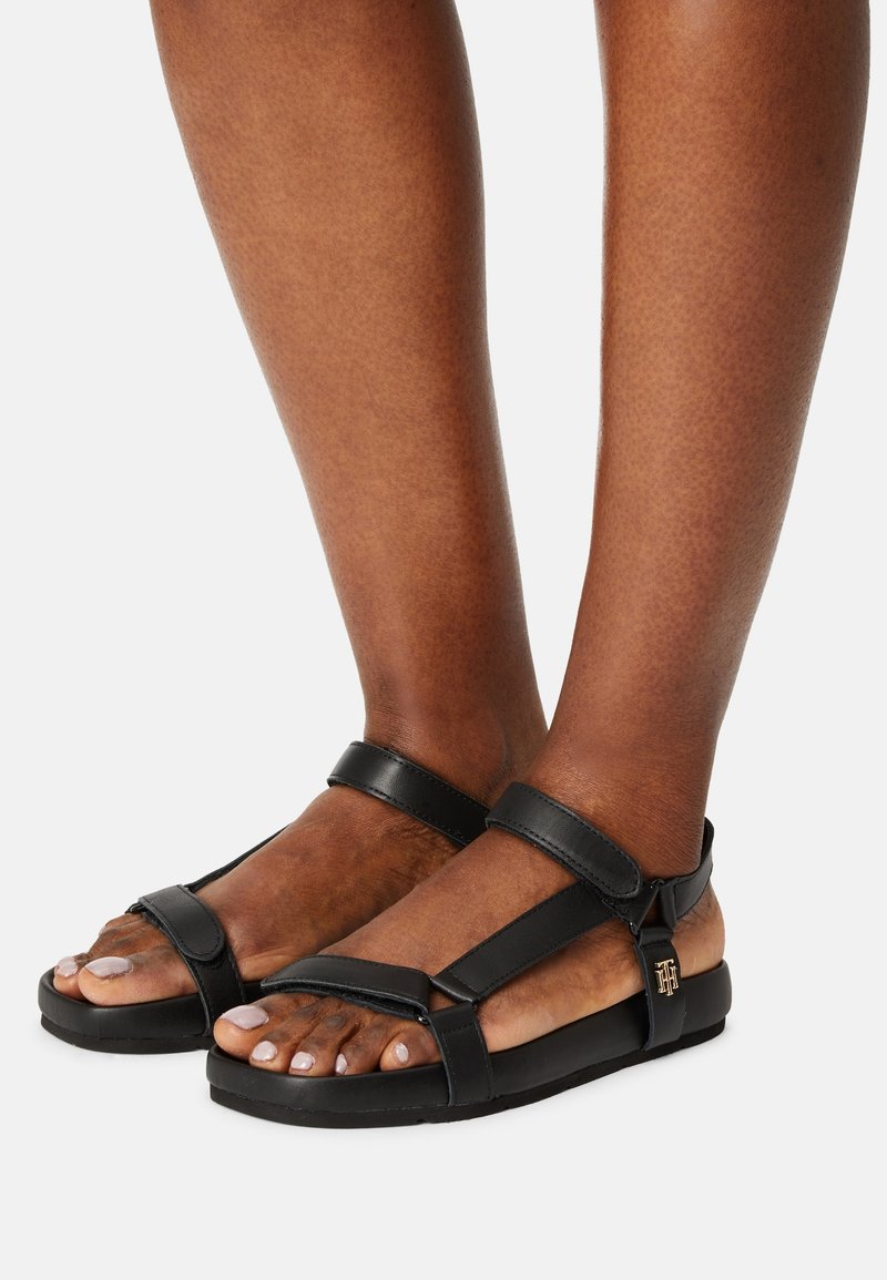 Pieds chaussés de sandales en cuir noir avec des sangles ajustables et un petit détail de logo doré sur le côté.