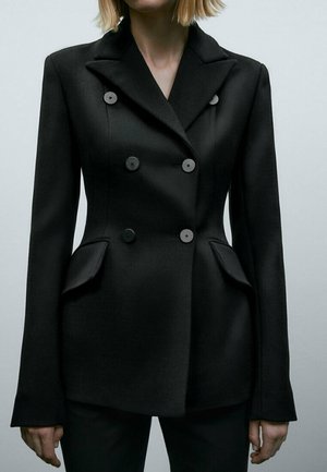 Femme portant un blazer noir cintré à double boutonnage avec revers à pointe et poches à rabat, debout devant un fond clair uni.