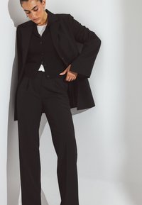 Črn krojen suknjič z dolgim plaščem in prilegajočim se žakardom ter belo srajco. Gladka tkanina, hlače ravnih hlačnic in široki rokavi na plašču.