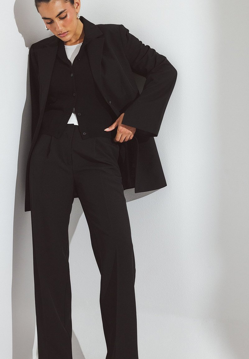 Črn krojen suknjič z dolgim plaščem in prilegajočim se žakardom ter belo srajco. Gladka tkanina, hlače ravnih hlačnic in široki rokavi na plašču.