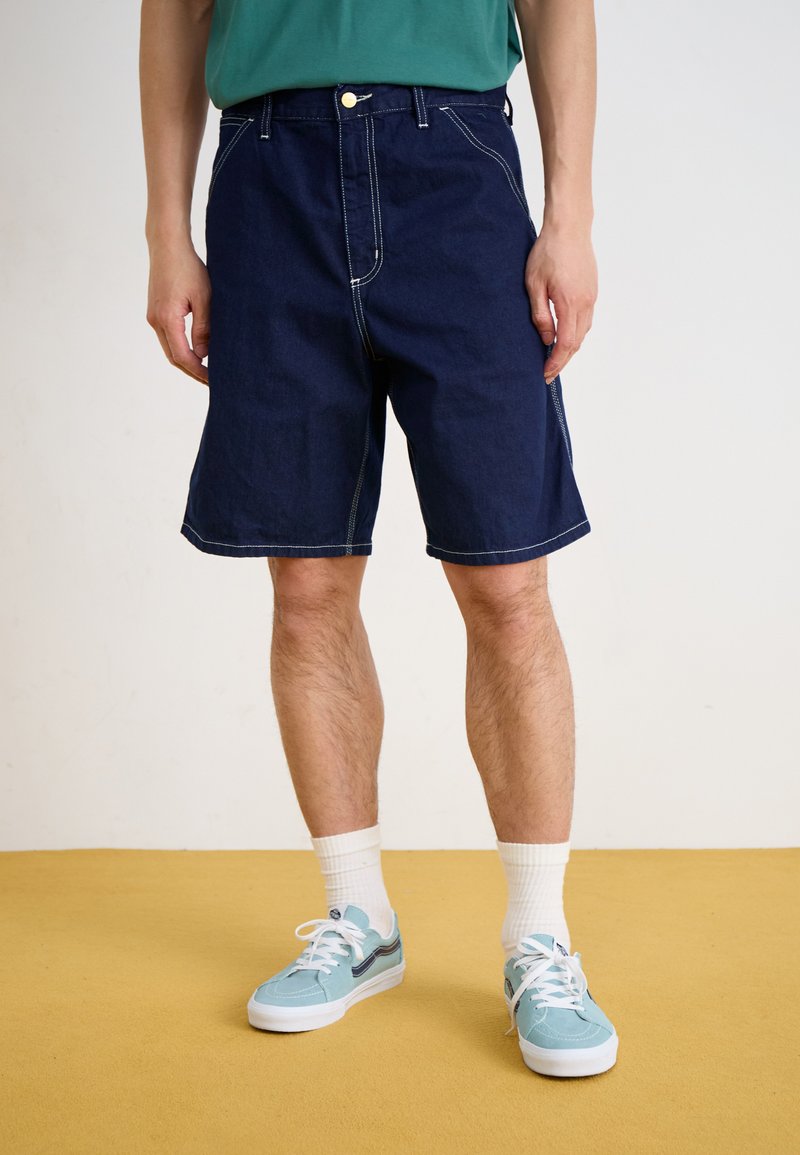 Pantalones cortos de mezclilla azul oscuro con un corte holgado y detalles de costura visibles. Combinados con zapatillas azul claro y calcetines blancos.