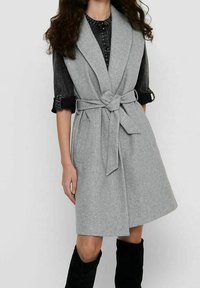 Cappotto senza maniche grigio chiaro in tessuto morbido con vita cintata, ampi revers e taglio al ginocchio, indossato sopra una camicia nera.