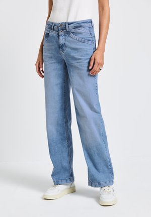 Jeans ampi di colore blu chiaro con una vita alta, dotati di chiusura con bottone e una leggera sfumatura. Abbinati a sneaker bianche.