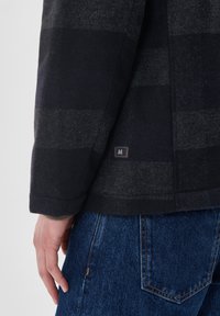 Chaqueta de mezcla de lana con un patrón de cuadros en gris oscuro y azul marino, con una pequeña etiqueta marcada como "M" en el dobladillo. Llevada sobre unos pantalones vaqueros azules.