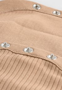 Dicker, gerippter Stoff in Tan mit silbernen Druckknöpfen. Nahaufnahme, die die Textur und die Details der Nähte sowie der Knopfbefestigungen hervorhebt.