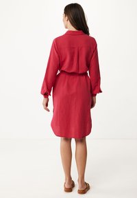 Röd linneklänning med långa ärmar, krage och cinchad midja. Har en ryggsöm och sidan slitsar. Bärs med bruna sandaler.