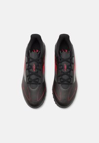 Svarta sportskor med röda accenter, med en texturerad övre del, snörning och en böjd sula. Horisontella ränder ger visuell detalj.