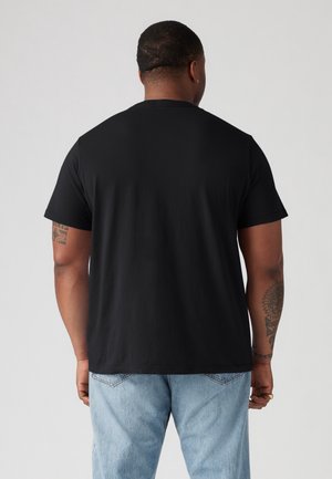 Mand iført en enkel sort T-shirt med korte ærmer og lyseblå jeans, set bagfra mod en ensfarvet lys baggrund.