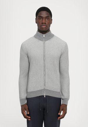 Man wearing a grey ribbed zip-up sweater with a high collar and dark pants, standing against a plain white background.