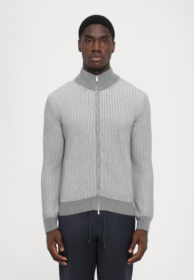Man wearing a grey ribbed zip-up sweater with a high collar and dark pants, standing against a plain white background.