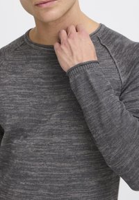 Chemise grise à manches longues en tissu tricoté texturé, avec un col rond et des poignets côtelés, présentant un motif marbré subtil.