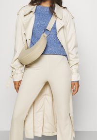 Trench-coat beige, pull en maille bleu, pantalon crème à côtes. L'accessoire est un sac crème incurvé avec une bandoulière beige. Design simple et moderne.