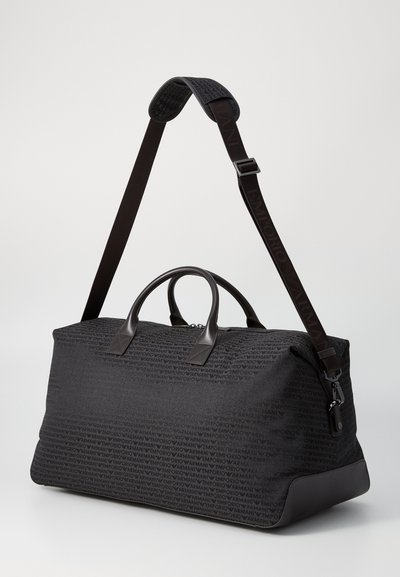 Sac de sport en tissu gris foncé avec un motif de logo répété, avec des finitions et des poignées en cuir, et un strap ajustable portant le logo.