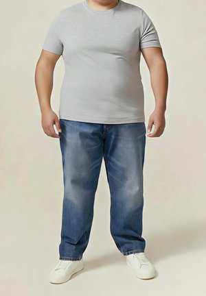 Hombre de pie, vistiendo una camiseta gris clara, vaqueros azules holgados y zapatos blancos sin cordones sobre un fondo beige liso.