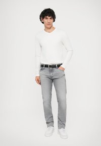 Graue Jeans mit schlanker Passform und leichtem Stretch, kombiniert mit einem enganliegenden weißen Langarmshirt. Weiße Sneaker und ein schwarzer Gürtel runden den Look ab.