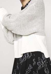 Maglione lavorato a maglia grigio e bianco con polsini e orlo a coste, dalla vestibilità ampia e superficie texturizzata. Abbinato a un vestito nero con figure astratte bianche.