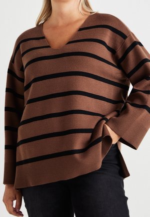 Femme portant un pull marron à col en V avec des rayures horizontales noires et un pantalon noir, posant avec une main dans la poche sur un fond blanc.
