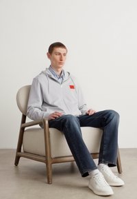 Lys grå hettejakke med rød logo-lapp, brukt over en blå rutete skjorte. Mørke jeans og hvite joggesko. Sitter på en stol med treframe.